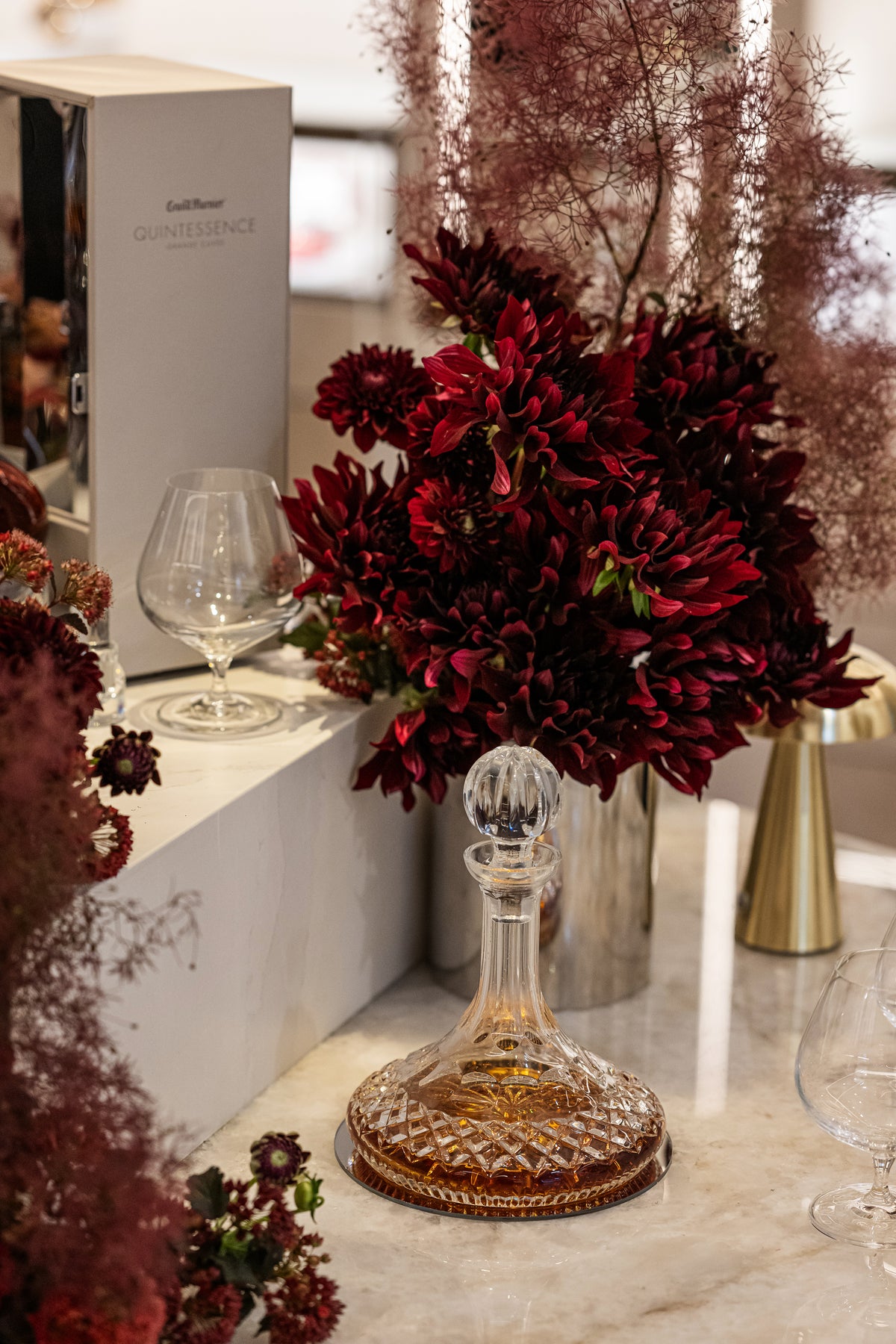 A cut-glass decanter of amber liquid beside deep red flowers and tasting glasses.