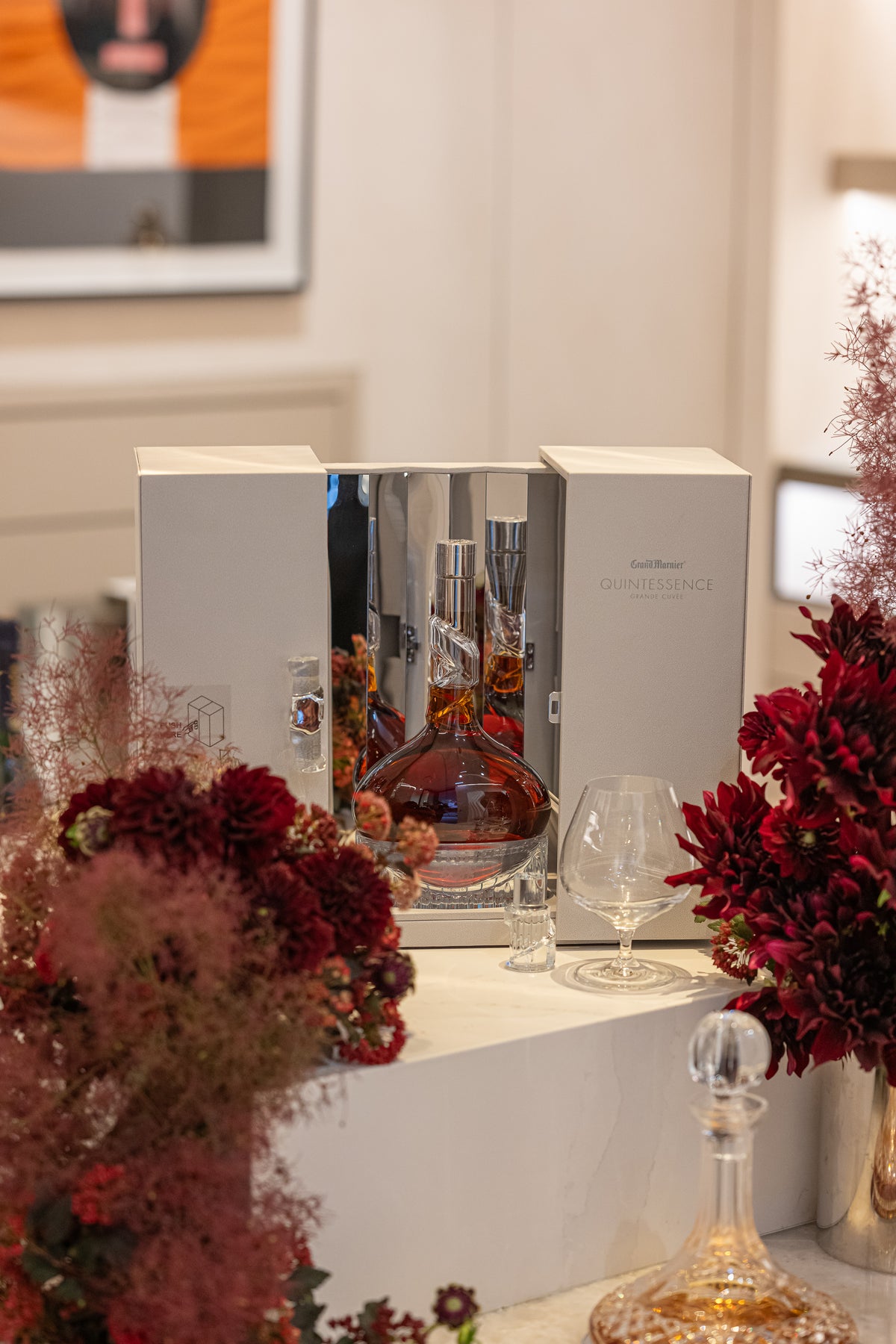 An amber-filled decanter in an open presentation box, framed by red flowers and tasting glasses.