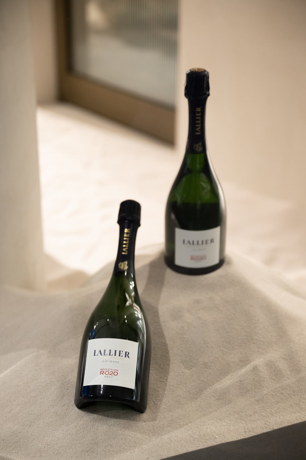 One green bottle standing behind another laid on pale draped fabric.