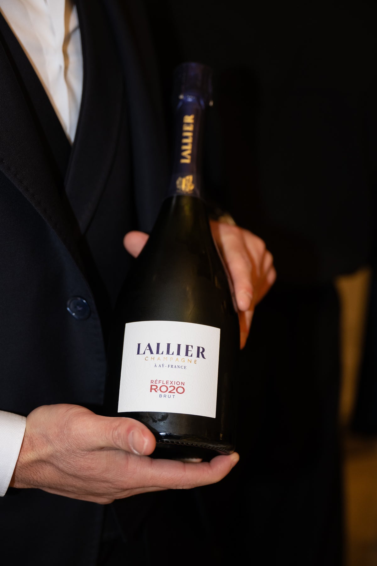 A person in a dark jacket holds a bottle with a white label towards the camera.