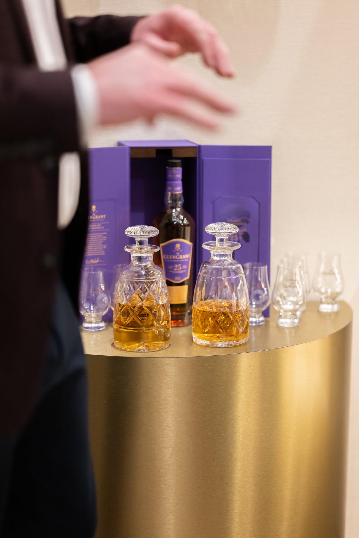 Two cut-glass decanters and tasting glasses on a round gold-coloured table in front of a purple presentation box.