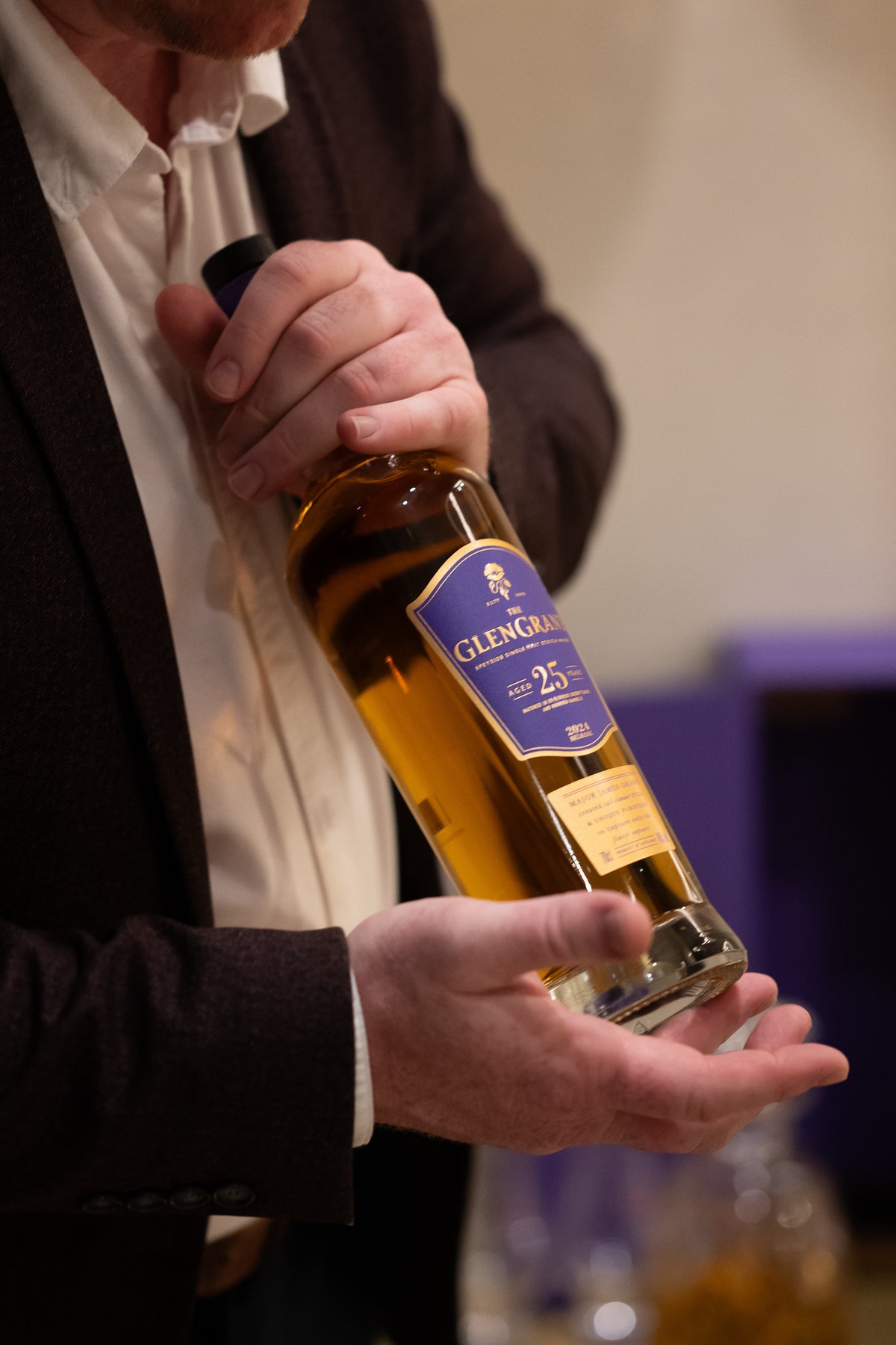 A person in a brown jacket holds an amber-filled bottle with a purple label.