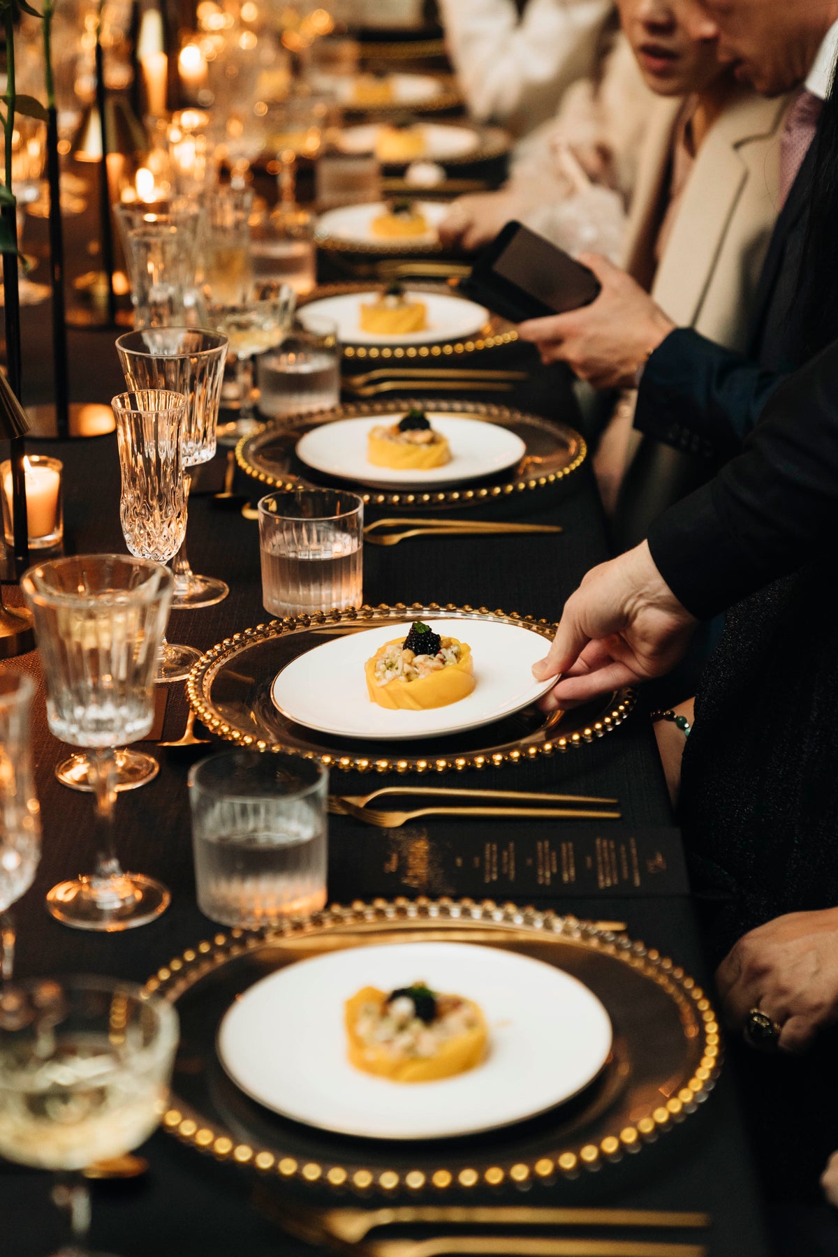 Hands placing white plates of food on a black dining table set with gold-edged plates and glassware.