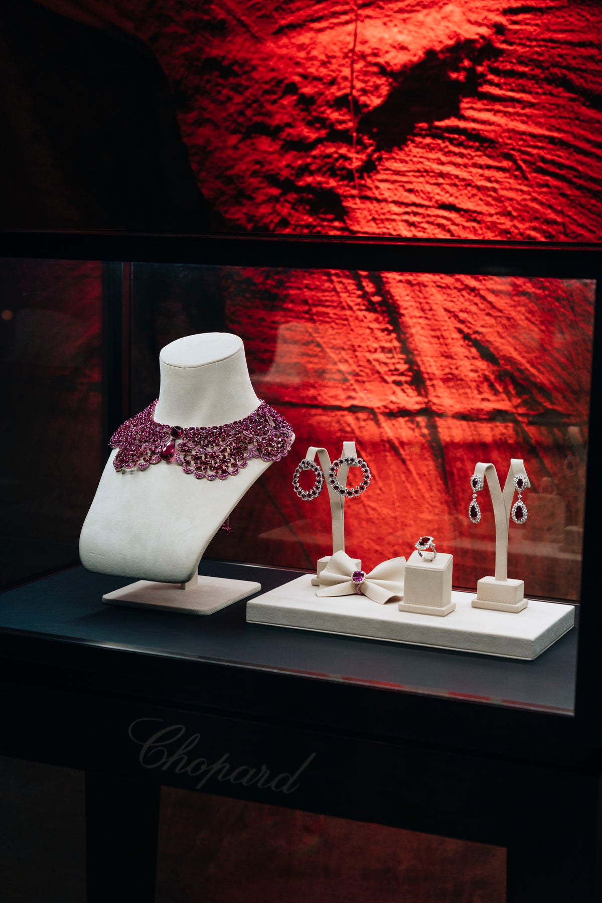 Pink floral necklace, earrings and rings displayed on white stands in a red-lit glass cabinet.