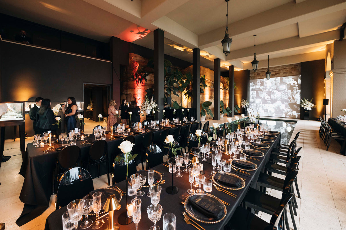 Long dining tables set with white roses and glassware beneath pendant lights in a warmly lit room.