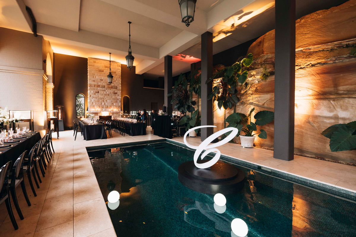 Pool with glowing white spheres and a large illuminated C, beside dining tables and a stone wall.