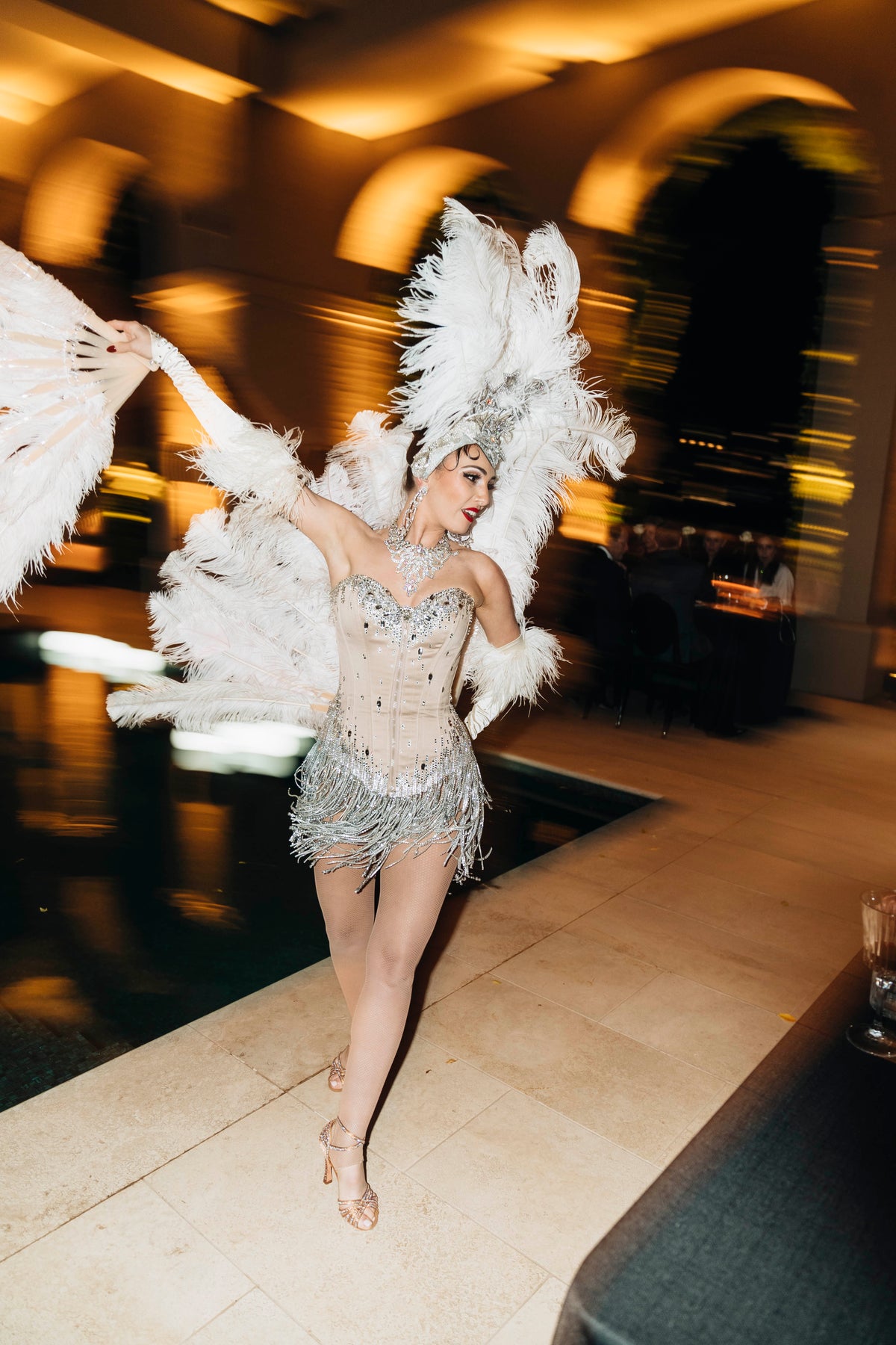 Performer in a sparkling silver costume and tall white feather headdress beside a pool.