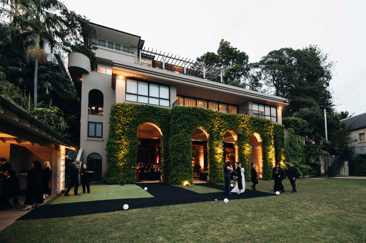 Vine-covered arches along a house, with guests standing on a black path across the lawn.