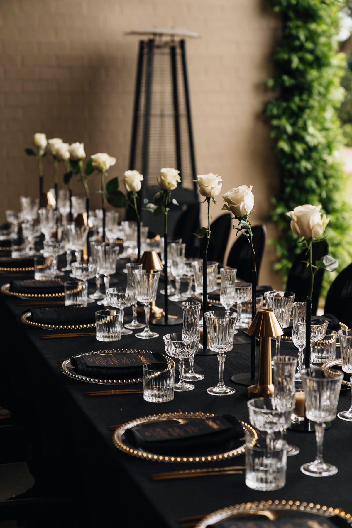 Long dining table dressed in black, with white roses, clear glassware and gold-edged plates.
