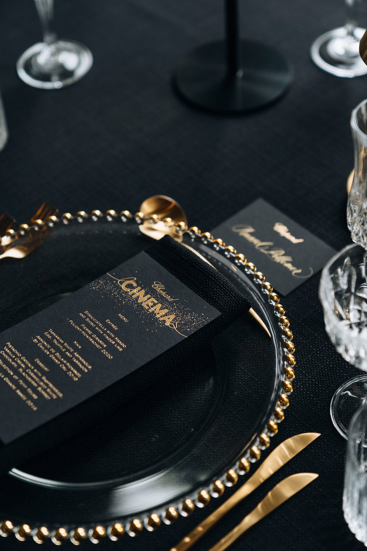 Black menu card on a gold-rimmed glass plate, with gold cutlery and clear glassware.