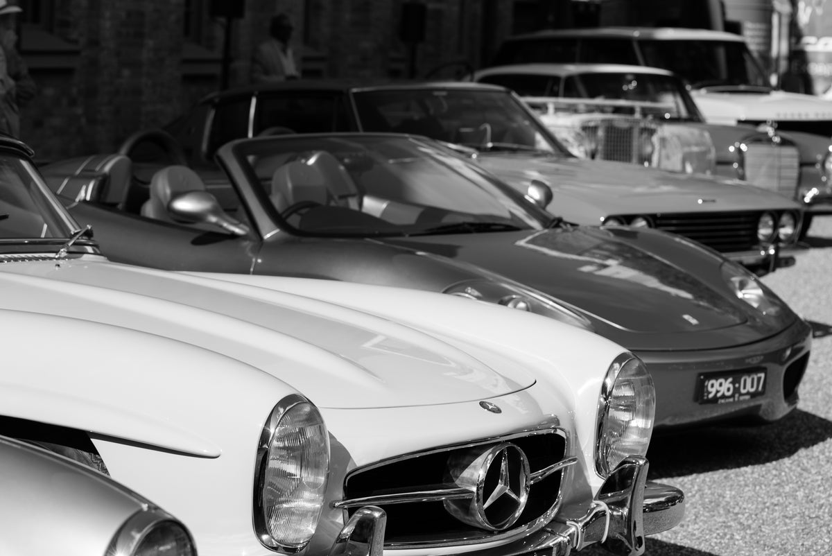 Black-and-white photograph of a white car's front grille, with other open-top cars parked behind.