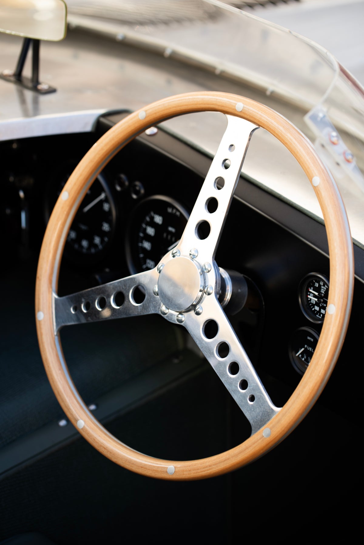 Wood-rimmed steering wheel with perforated metal spokes in front of a black instrument panel.