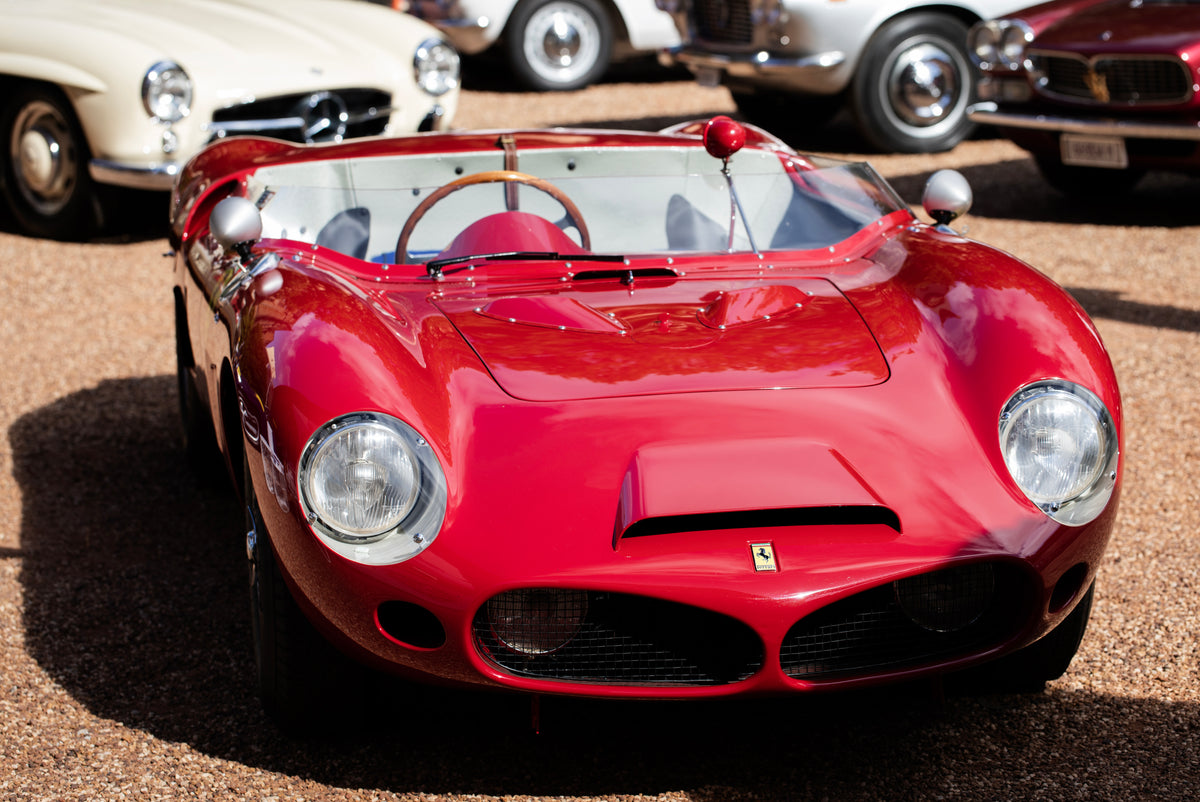 Red open-top sports car with round headlights and a small windscreen, parked on gravel.