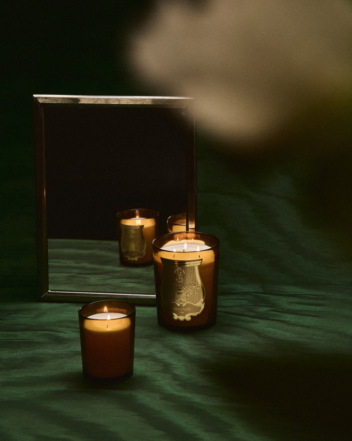 Lit Trudon candles reflected in a mirror on deep green fabric